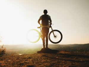 A cyclist stands silhouetted with a mountain bike on a hilltop during sunset, evoking adventure and freedom.