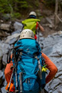 Two hikers with backpacks and helmets navigating a rocky trail in the forest.