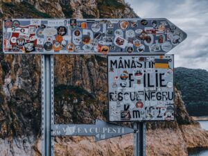 Direction signs with various stickers located on roadside near rocky cliff in sea against cloudy sky in Romania