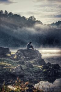 A lone figure reflects on a rock by a misty lake in Rancabali amidst tranquil surroundings.