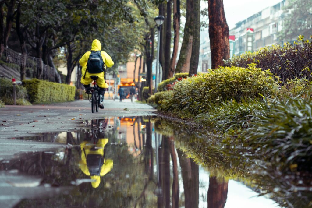A person in a yellow raincoat cycles along a wet street with reflections in a puddle.